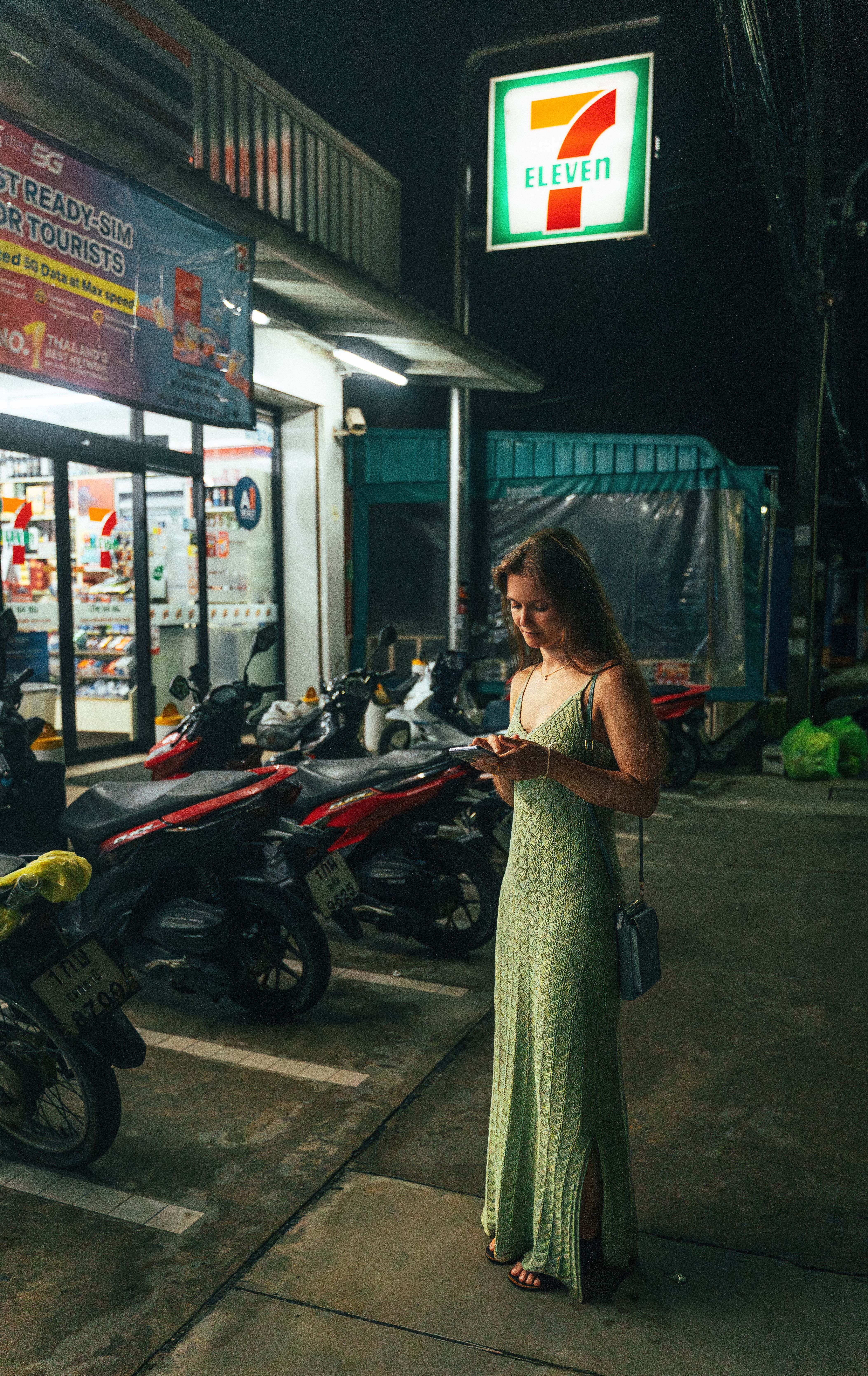Woman with smartphone standing outside a 7-Eleven in Phuket at night.