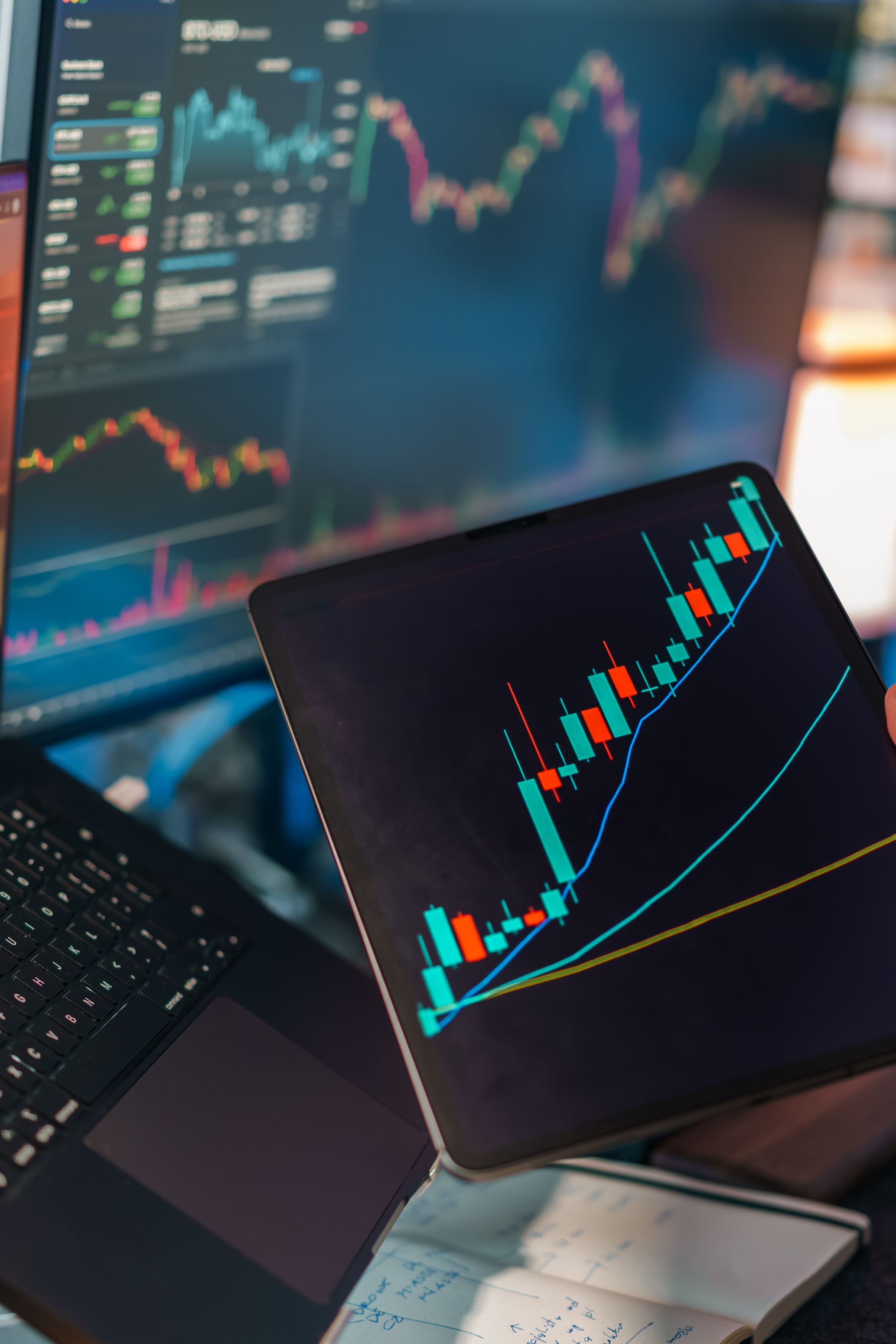 A tablet displaying a candlestick chart with a laptop screen showing stock market data for financial analysis.