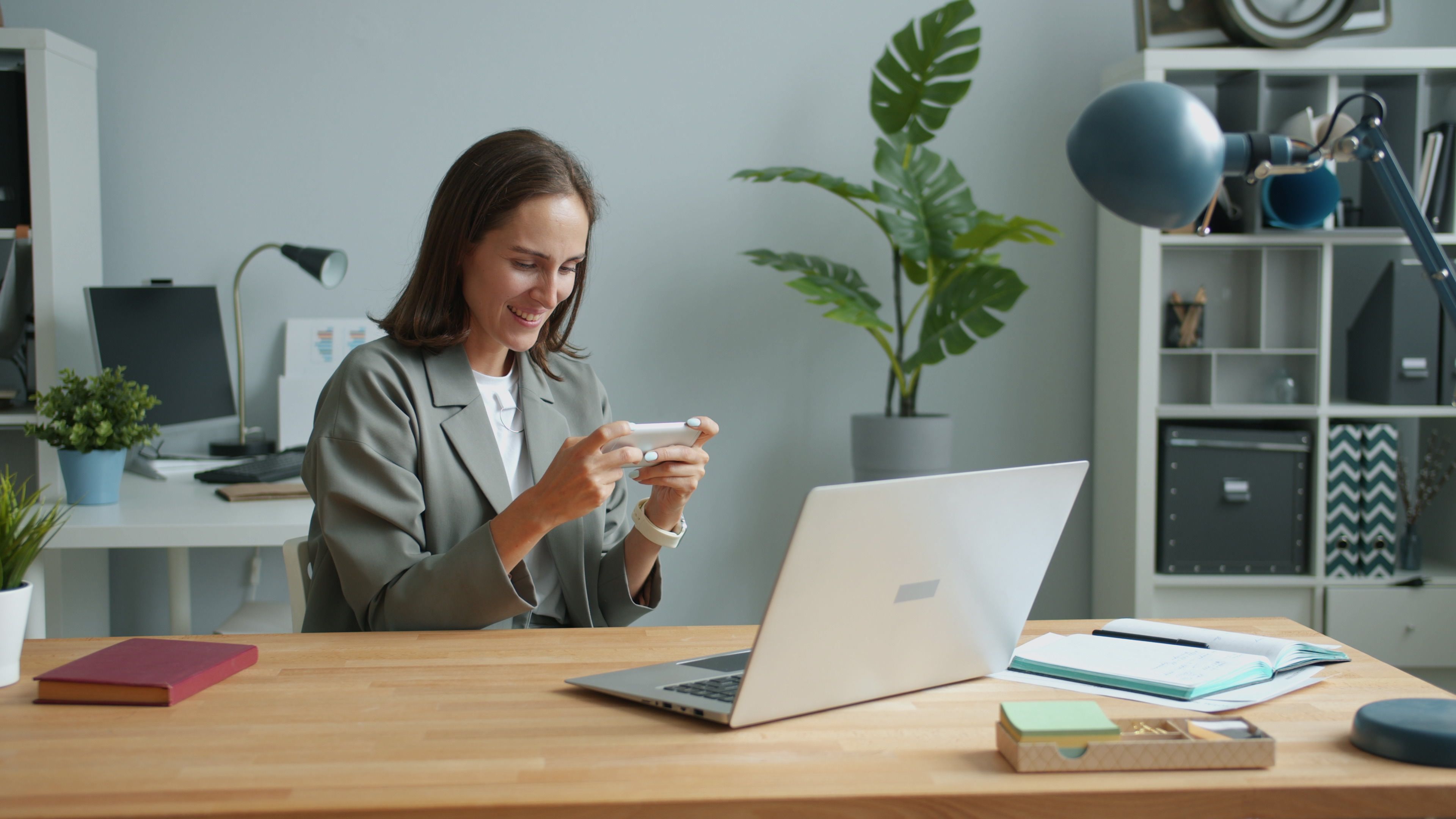 A woman in a home office using a smartphone while working on a laptop.