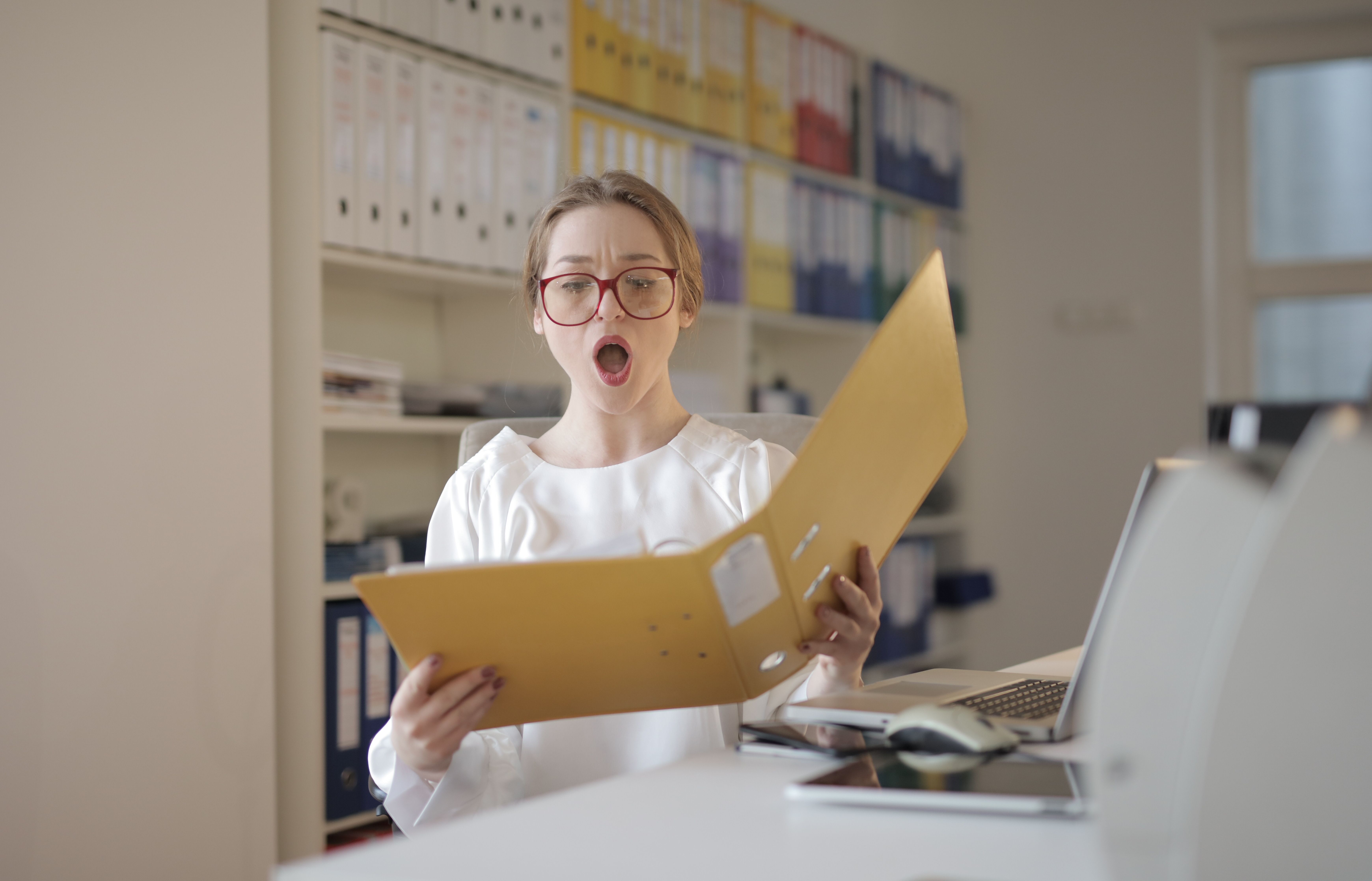 Surprised woman with eyeglasses reading a file in an office filled with shelves of records.
