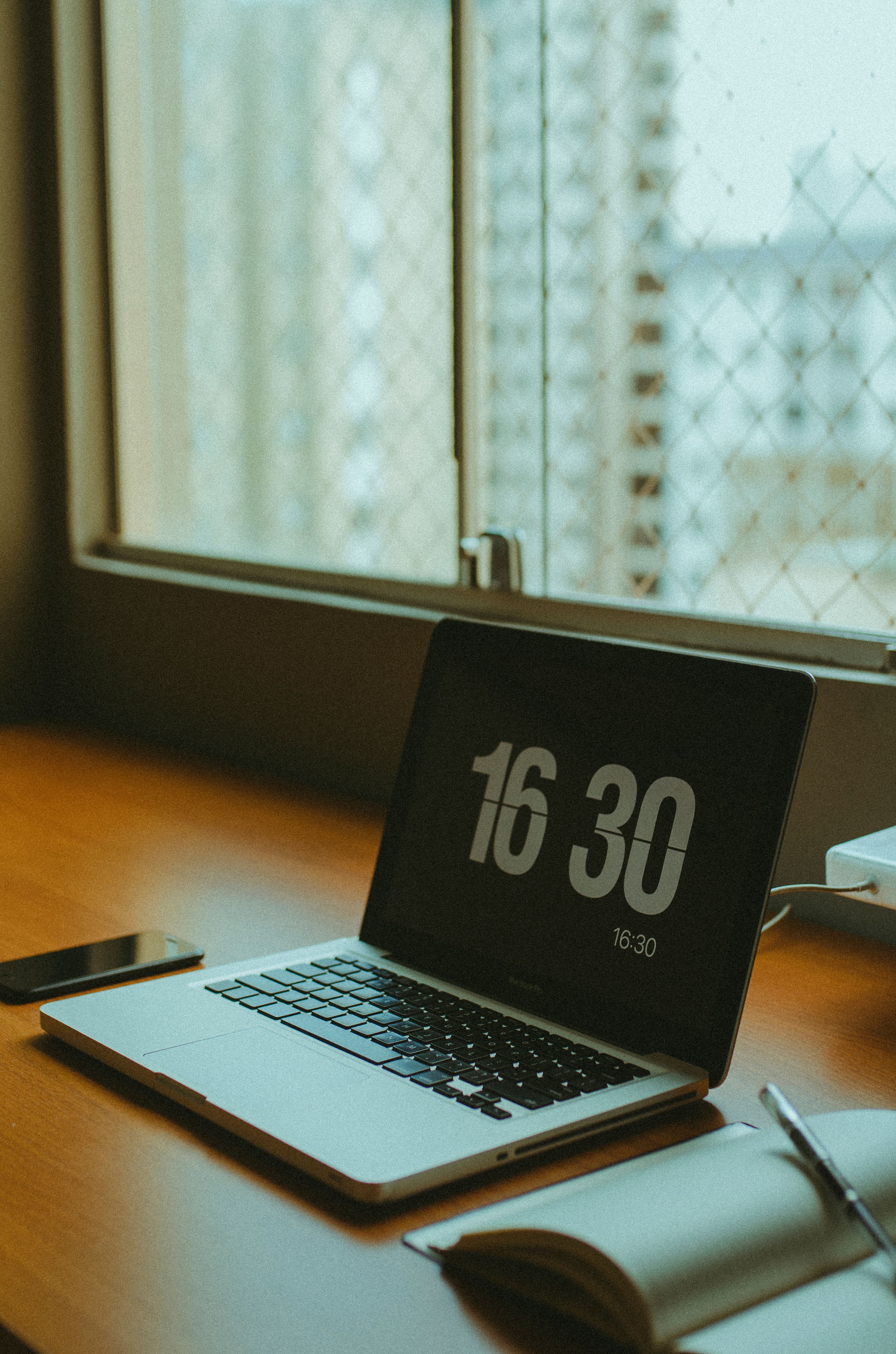 A sleek workspace featuring a laptop, smartphone, and notebook by a window.